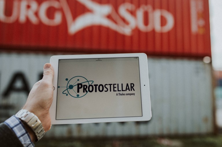 Eine Männerhand hält ein Tablet auf dem das Wort Protostellar geschrieben steht. Im Hintergrund sind Container zu sehen.